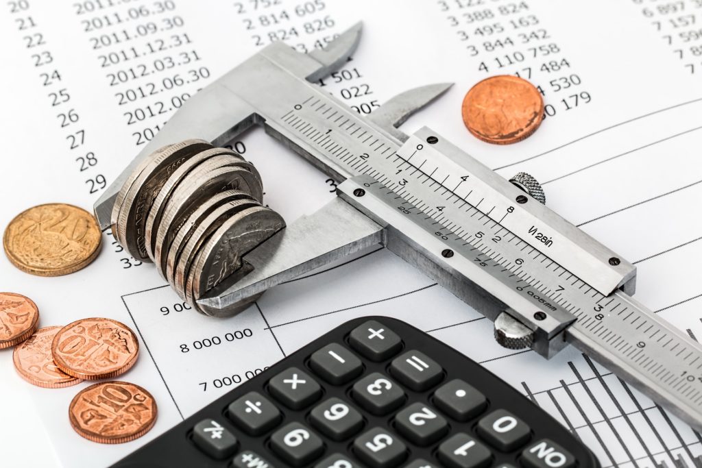Image of caliper holding a stack of coins next to a calculator. The caliper is laying on a budget report.