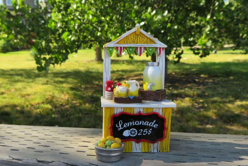 Image of a lemonade stand. Stand consists of a wooden booth painted white and yellow stripes. A painted sign says "Lemonade .25 cents". There is a full pitcher of lemonade and red cups.