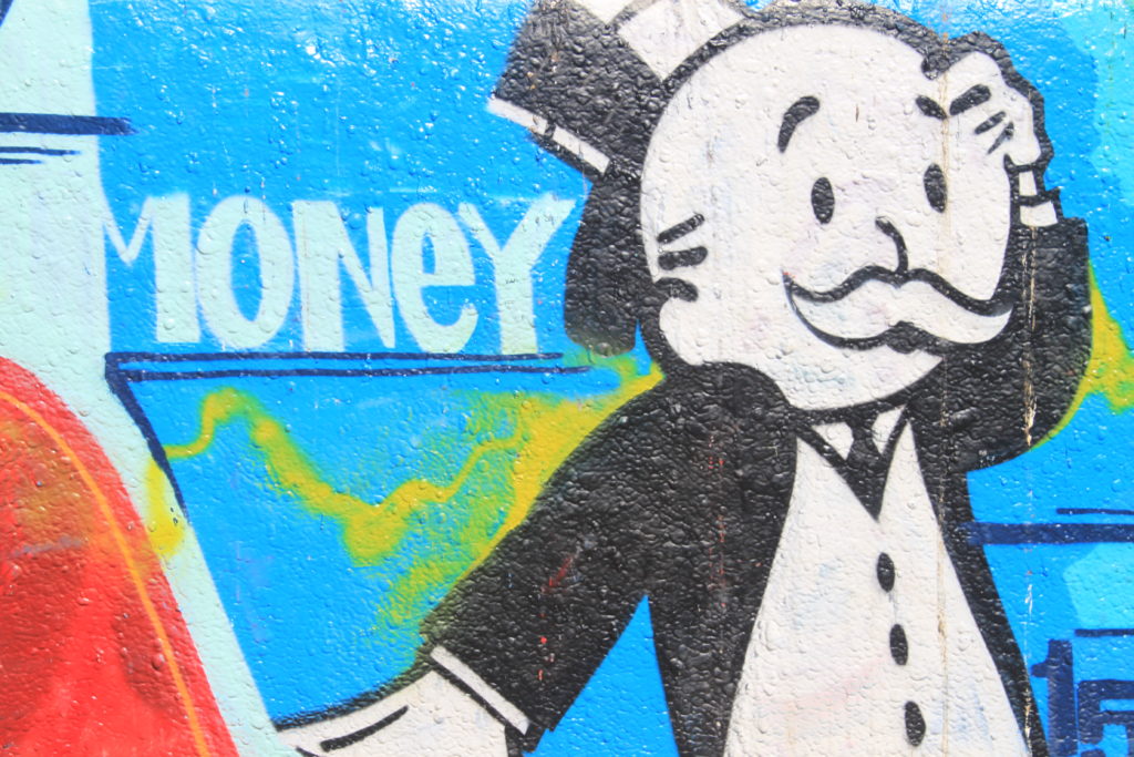 Image of street art showing the main male character from the game Monopoly. The character wears a black top hat and an old fashioned tuxedo. The character has a worried look on their face. In the background is the word "Money" in white letters.