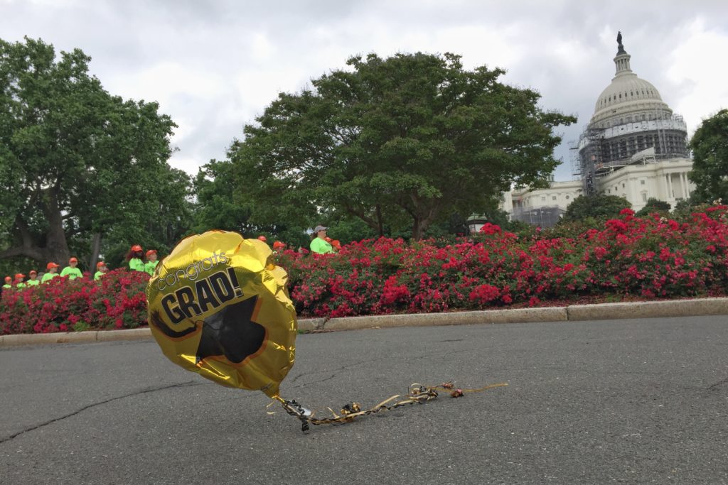 Image of a deflated gold balloon with the text "Congrats Grad!". In the near background are trees and rose bushes. In the far background is the United States of America capitol congress building.