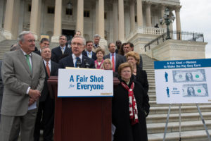 Image of group of United States government officials with one speaker at a podium. On the podium there is a sign with the text "A fair shot for everyone". Another sign to the right of the podium says "A fair shot for women to make the pay the earn". The second sign also has the images of two dollar bills and the silhouettes of a man and woman. The dollar bill next to the woman is smaller and has the text "seventy seven cents for every dollar a man makes".
