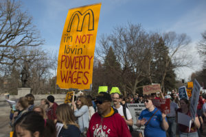 Image of a group of people gathered for a strike march. People have hand painted signs. Most prominent sign has a yellow background, the "M" logo from McDonals, and the text "I'm not lovin' poverty wages"