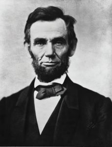 Black and white photo of Abraham Lincoln. He has a straight face and wears a suit and bowtie.