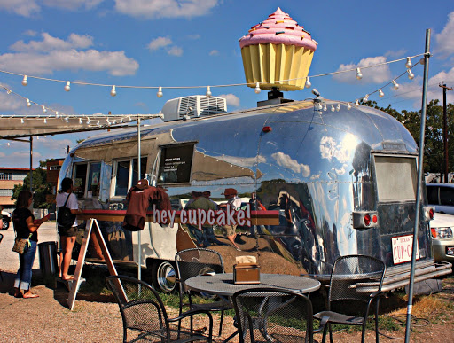 Silver foodtruck parked with an awning out. The side of the truck reads "Hey Cupcake!" and the van has a huge cupcake on top of it.