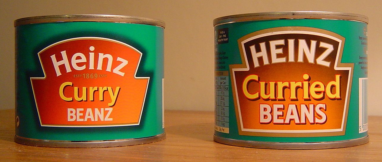 Two cans of Heinz baked beans sit on a table. The first: Curry Beanz. Second: Curried Beans.