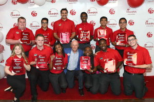 Twelve chick-fil-a employees pose for a group picture in their red uniforms while holding giant sauces with their names on them.