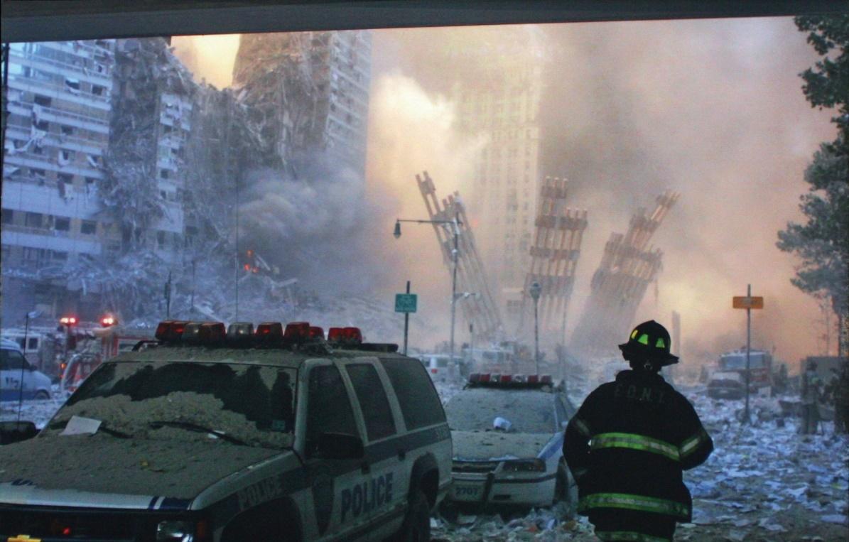 firefighter in the foreground with two broken down emergency vehicles; wreckage of buildings in the 9/11 attacks in the background. 