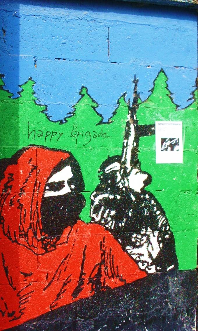 mural with a person in a black mask and a red hood against a background of green trees and blue sky; "happy brigade" is  scrawled at the top.