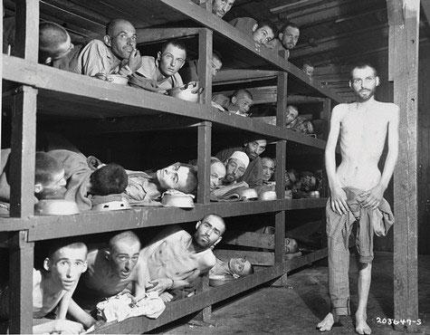 image of emaciated men laying crowded in  wooden bunks stacked four high. One man is standing, leaning against a vertical wooden post.