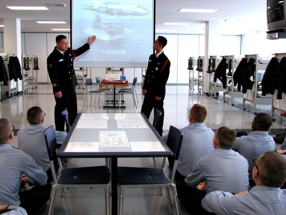 Un hombre de uniforme dando una presentación a un pequeño grupo de aprendices militares