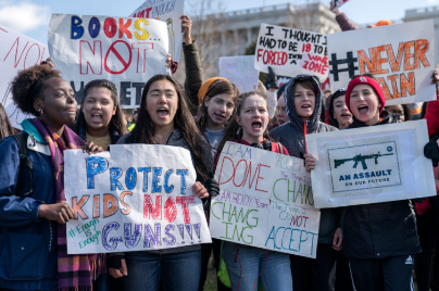 Imagen de grupo de jóvenes sosteniendo carteles en un mitin político. Las señales dicen mensajes como: “¡Proteja a los niños no a las armas # Ya basta” y “Libros no balas” y “# nunca más” y un letrero con una imagen de un fusil de asalto con el texto “Un asalto a nuestro futuro”.