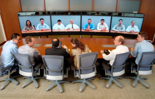Una reunión en la que seis miembros están presentes juntos en una sala de conferencias, y ocho miembros se unen a través de videoconferencia.