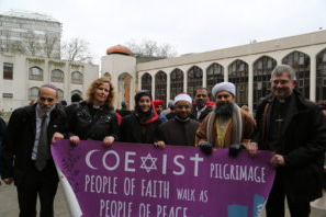 Foto de seis personas de diferentes religiosos sosteniendo un cartel morado con texto en blanco. El letrero dice “Conexistir peregrinación gente de fe camina como gente de paz”.