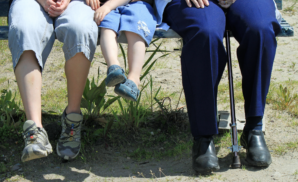 Three people of different ages sitting on a bench. The person on the left is in the 30-40 year-old range, the person in the middle is in the 5-10 year old range, and the person on the left has a cane and is in the 60-75 year old range.