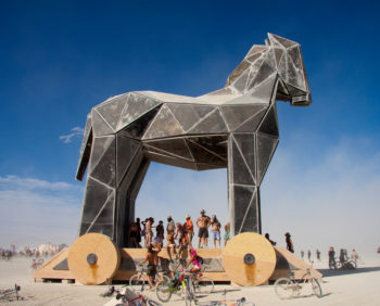 Photo of a large Trojan horse outside. People stand around at the base of the horse.