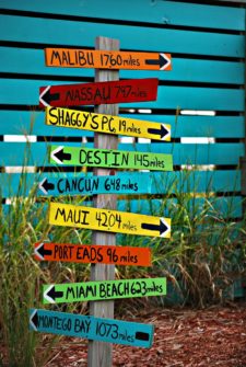 A sign post with nine different signs pointing to different locations with the distance to each location written out. Malibu is 1760 miles away. Nassau is 797 miles away. Shaggy's PC is 19 miles away. Destin is 145 miles away. Cancun is 648 miles away. Maui is 4204 miles away. Port Eads is 96 miles away. Miami Beach is 626 miles away. Montego Bay is 1073 miles away.