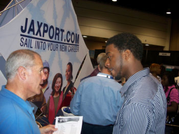 Un hombre habla con otro, hombre más joven en una feria de trabajo. Al fondo se encuentra un cartel publicitario de Jaxport.com. “Navega hacia tu nueva carrera”.