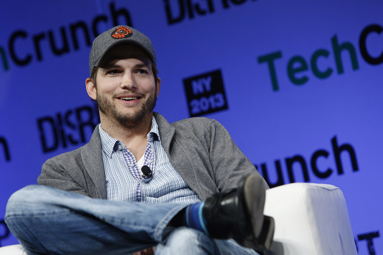 Uma fotografia mostra Ashton Kutcher sentado em frente a uma tela digital que diz Tech Crunch, N Y 2013.