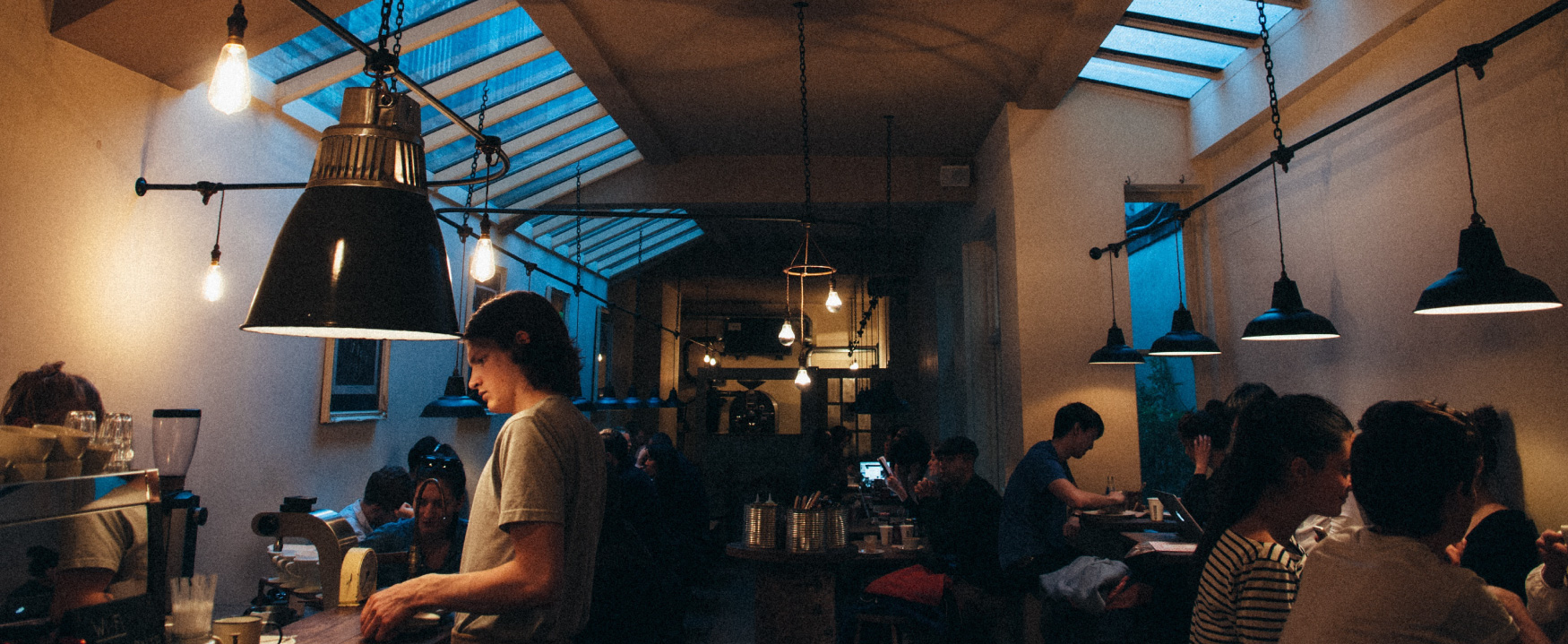 Uma cafeteria mal iluminada e lotada é mostrada. Os clientes se sentam às mesas e um trabalhador atende um cliente no balcão.