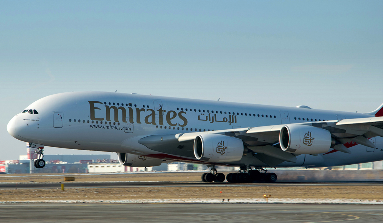 Une photographie montre un grand avion à deux étages, avec le mot « Emirates » peint sur le côté. Il y a aussi du sanskrit peint sur le flanc de l'avion.