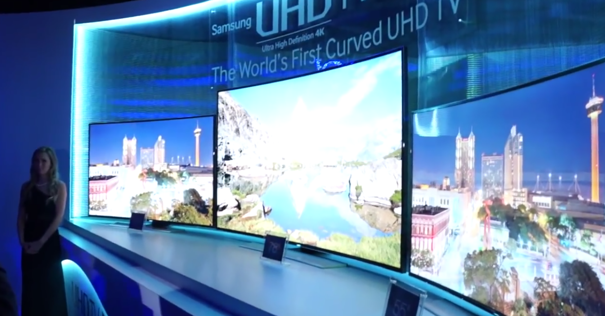 一张照片显示了一台带有凹形屏幕的大型电视。 电视显示屏上方有一个标语，上面写着 Samsung U H D，世界上第一台曲面的 U H D T V