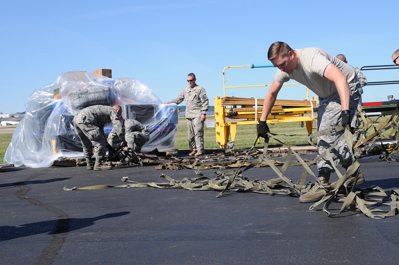 A photograph shows a group of men wearing military fatigues, using heavy duty strapping to secure a large pallet stacked with cargo.