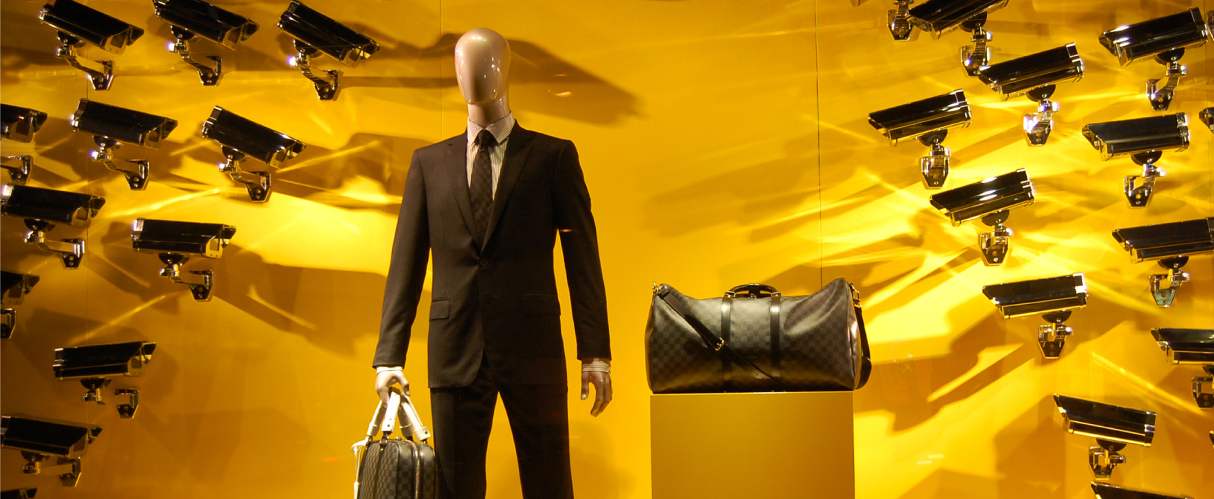 A display shows a mannequin dressed in a business suit, holding a designer bag, with a second designer bag on a pedestal beside it. On the wall behind the mannequin are dozens of security cameras pointed at it, and the bags.