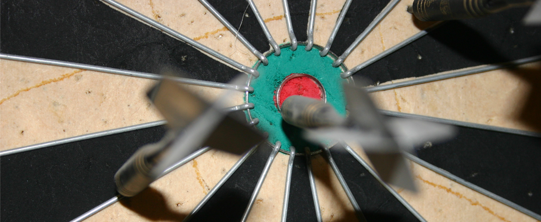A photograph shows a dart board with a few darts stuck to the board, on is sticking into the bullseye.