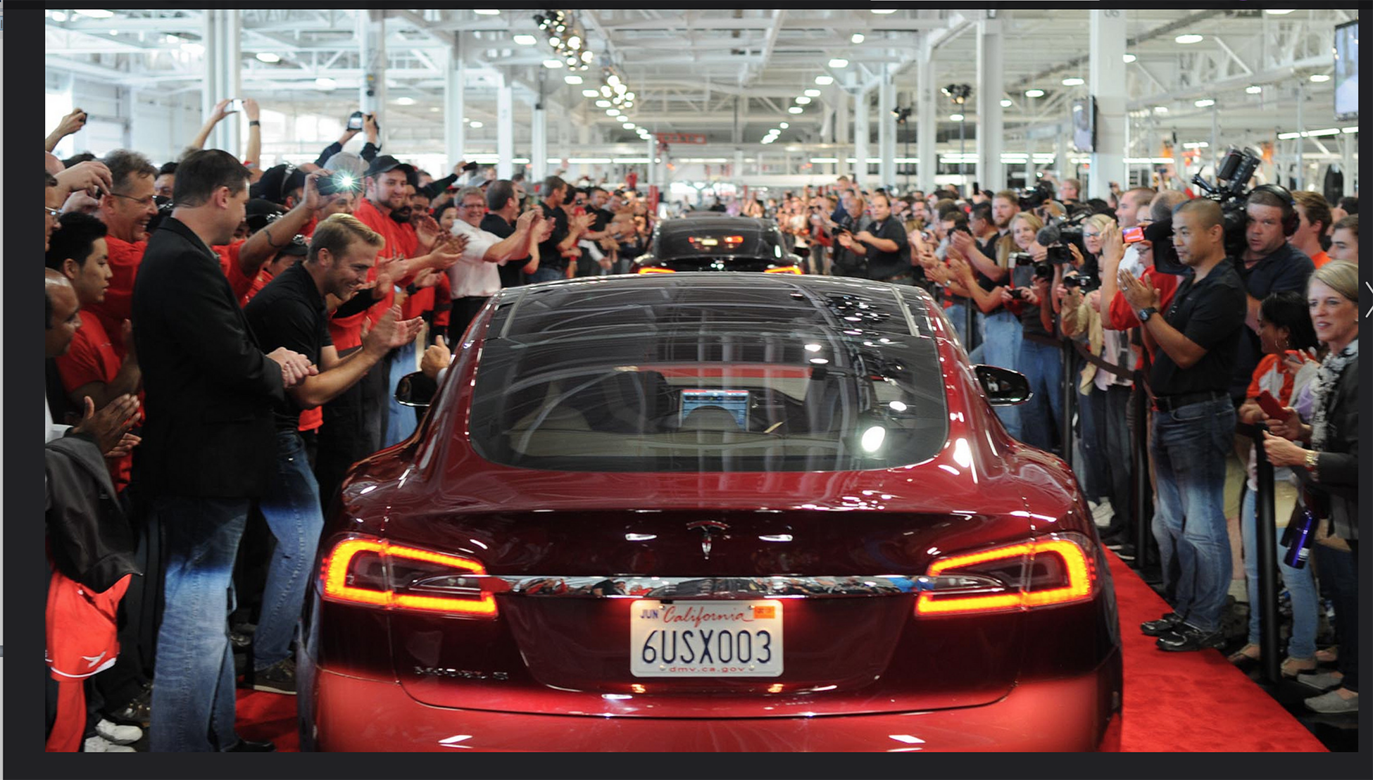 Une photographie montre une foule nombreuse encourageant une file de voitures Tesla roulant sur un tapis rouge.