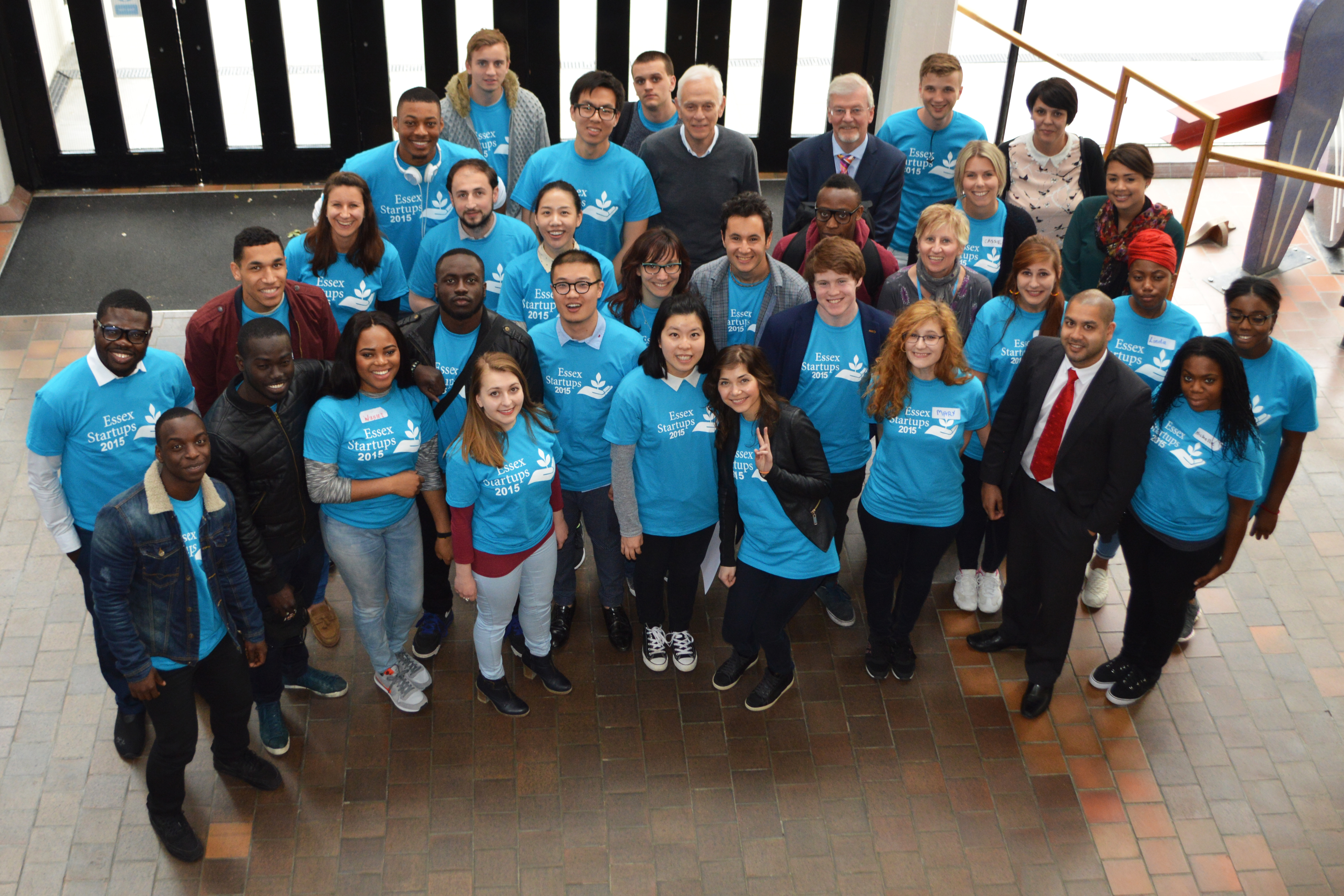 A photograph shows a group of people, made up of younger and older people, wearing shirts that read, Essex Startups 20 15.
