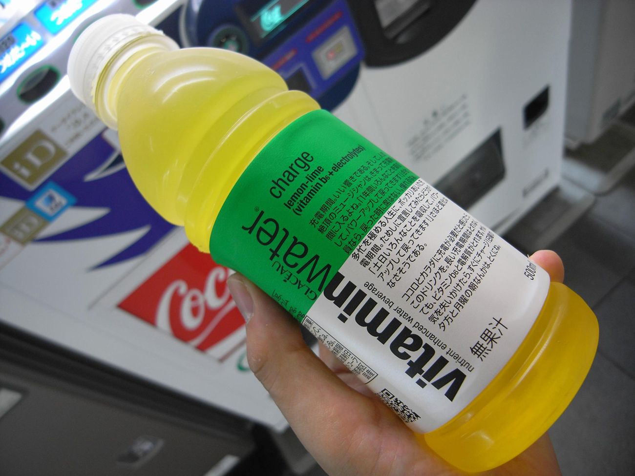 A photograph shows a person holding a bottle of Vitamin water. The label is written in both English and Japanese lettering.