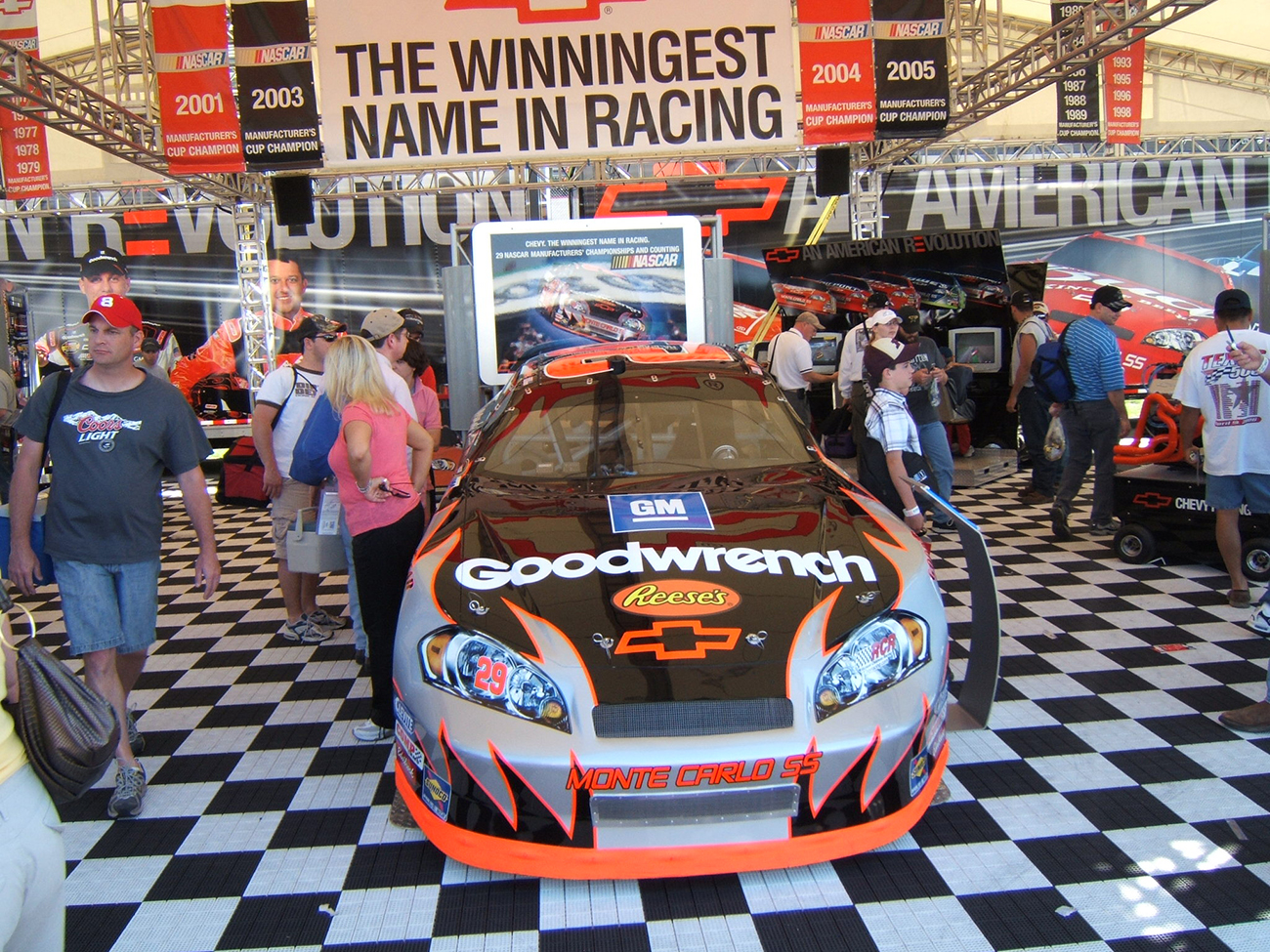 A photograph shows a group of people walking around a Nascar display area. There is a large Chevy racecar parked inside, covered in decals such as G M, Reese's candy, and Good wrench.