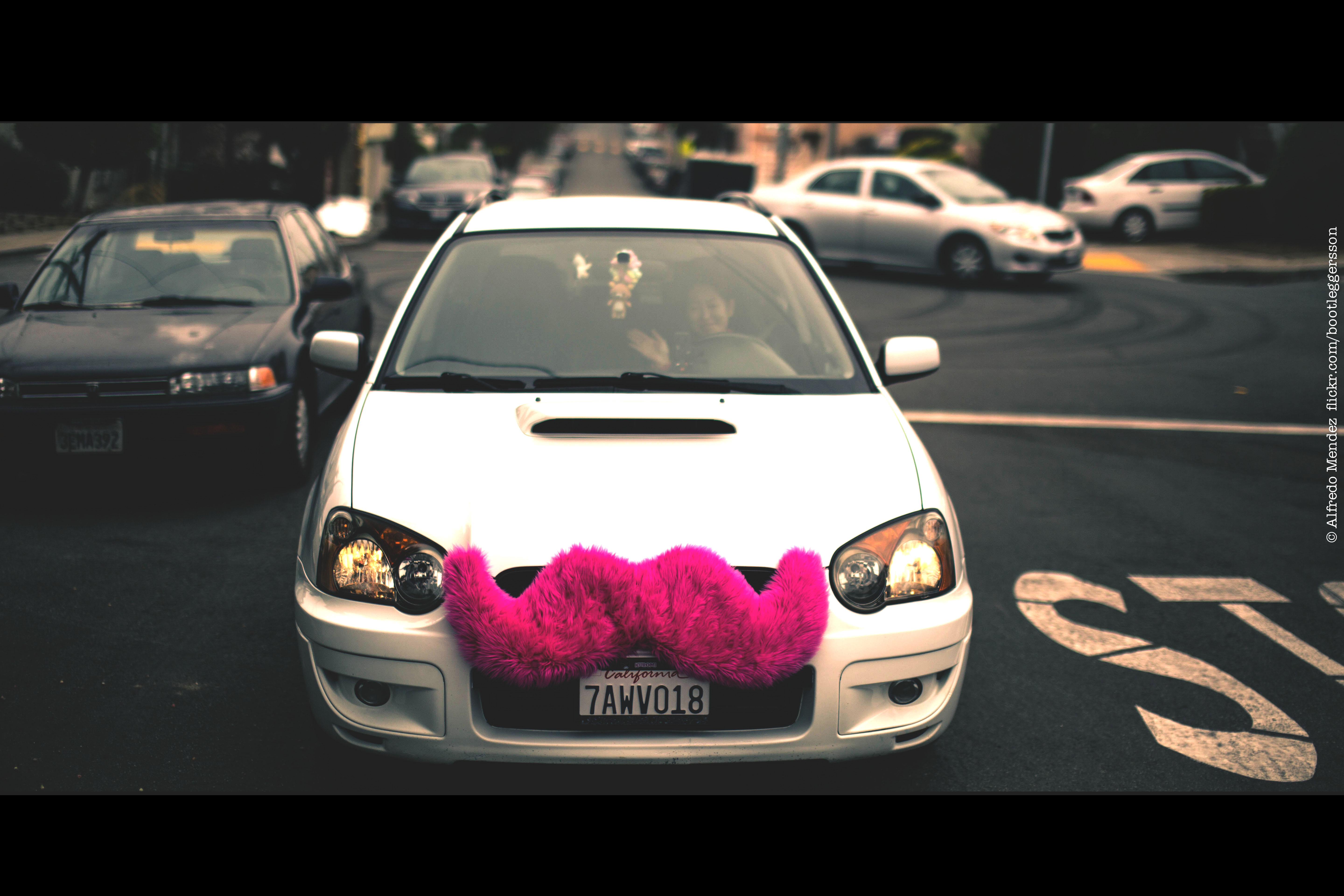 Une photographie montre une voiture avec une grande moustache rose duveteuse fixée à la calandre avant de la voiture.