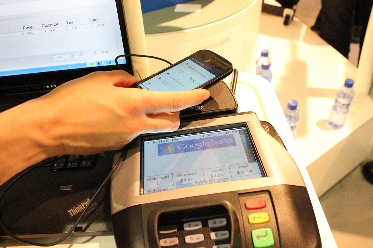 Uma foto mostra uma máquina de ponto de venda em uma loja. A tela da máquina diz: Google Wallet e uma pessoa está segurando o telefone acima da base plana ao lado da tela.
