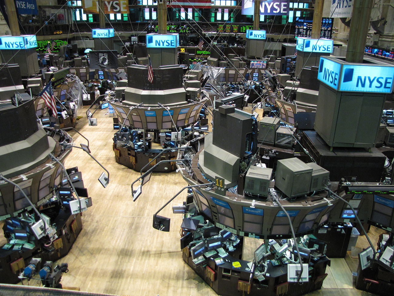 تظهر صورة بورصة نيويورك. تحتوي المنطقة الداخلية على العديد من المحطات، كل منها مغطى بأجهزة الكمبيوتر وشاشات العرض. توجد في الخلفية شاشات تعرض الأسهم والأسعار.