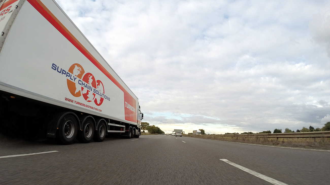 照片显示一辆大型货运卡车在高速公路上行驶。 卡车侧面的徽标写着 “供应链解决方案”；w w w dot turner dash distribution dot com。