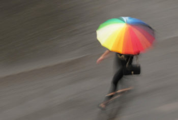 Foto de una mujer corriendo bajo la lluvia sosteniendo un paraguas de color arcoíris.