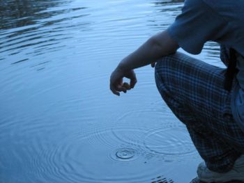 Photo of someone crouching beside a pond, apparently having just dropped a stone into the water. Concentric circles can be seen where the stone dropped in.