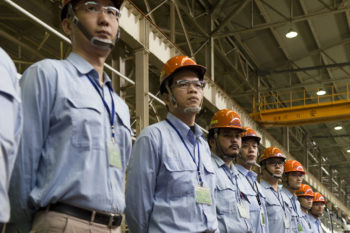 Long row of identically dressed Chinese workers. Each wears a red hard hat, pressed blue shirt and ID tag, khaki pants, and black belt.