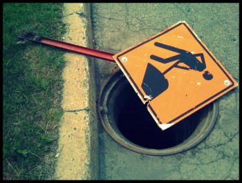 Photo of a hole in street (without its manhole cover), partially covered by a "men at work" street sign. Photo tag: "lawsuit waiting to happen."