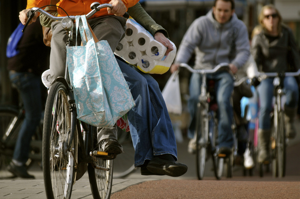 Photo of the lower half of a couple riding a bike. The person in the back sits sidesaddle and is carrying a large package of toilet paper.