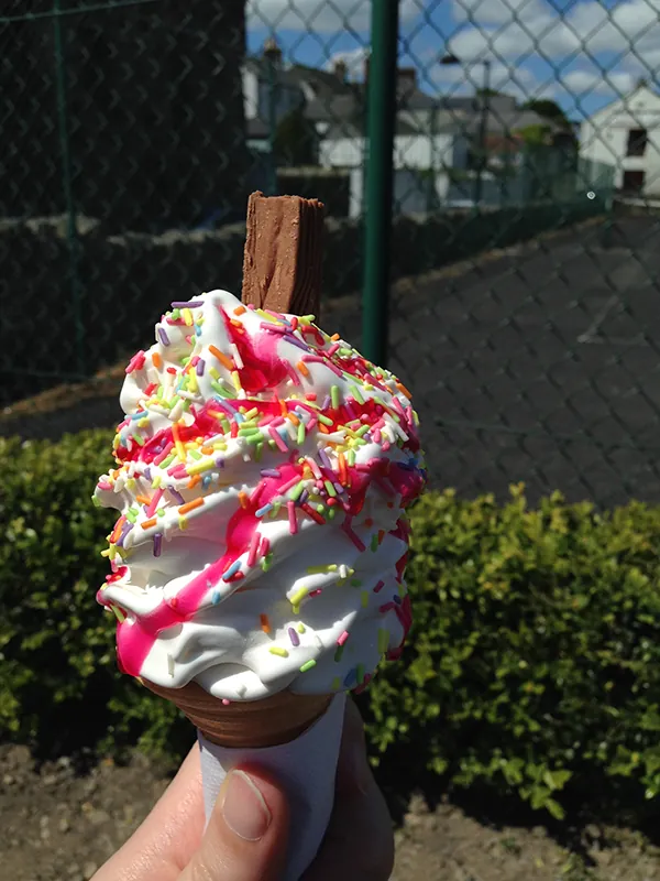 A hand holds a vanilla ice cream cone with strawberry syrup and rainbow sprinkles.