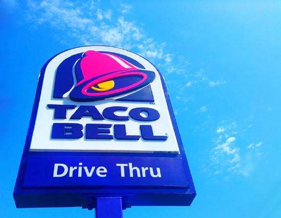 Image of a Taco Bell drive through sign is seen with blue sky and clouds behind it.