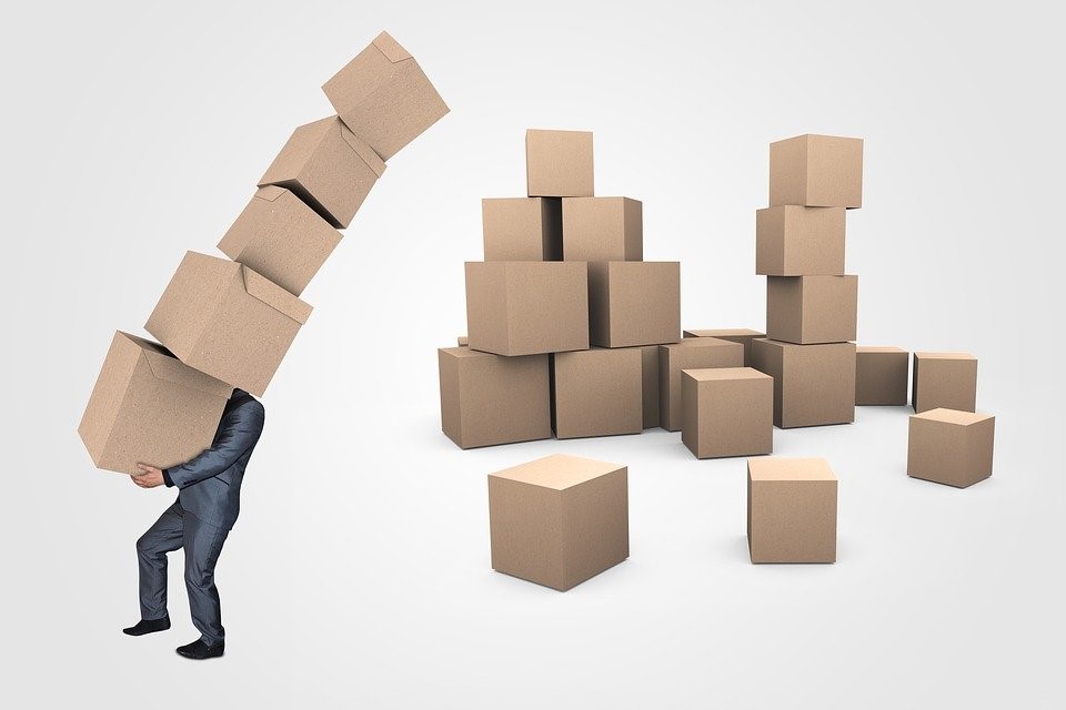 A worker trying to move a stack of boxes taller than he is. Other boxes are stacked nearby.