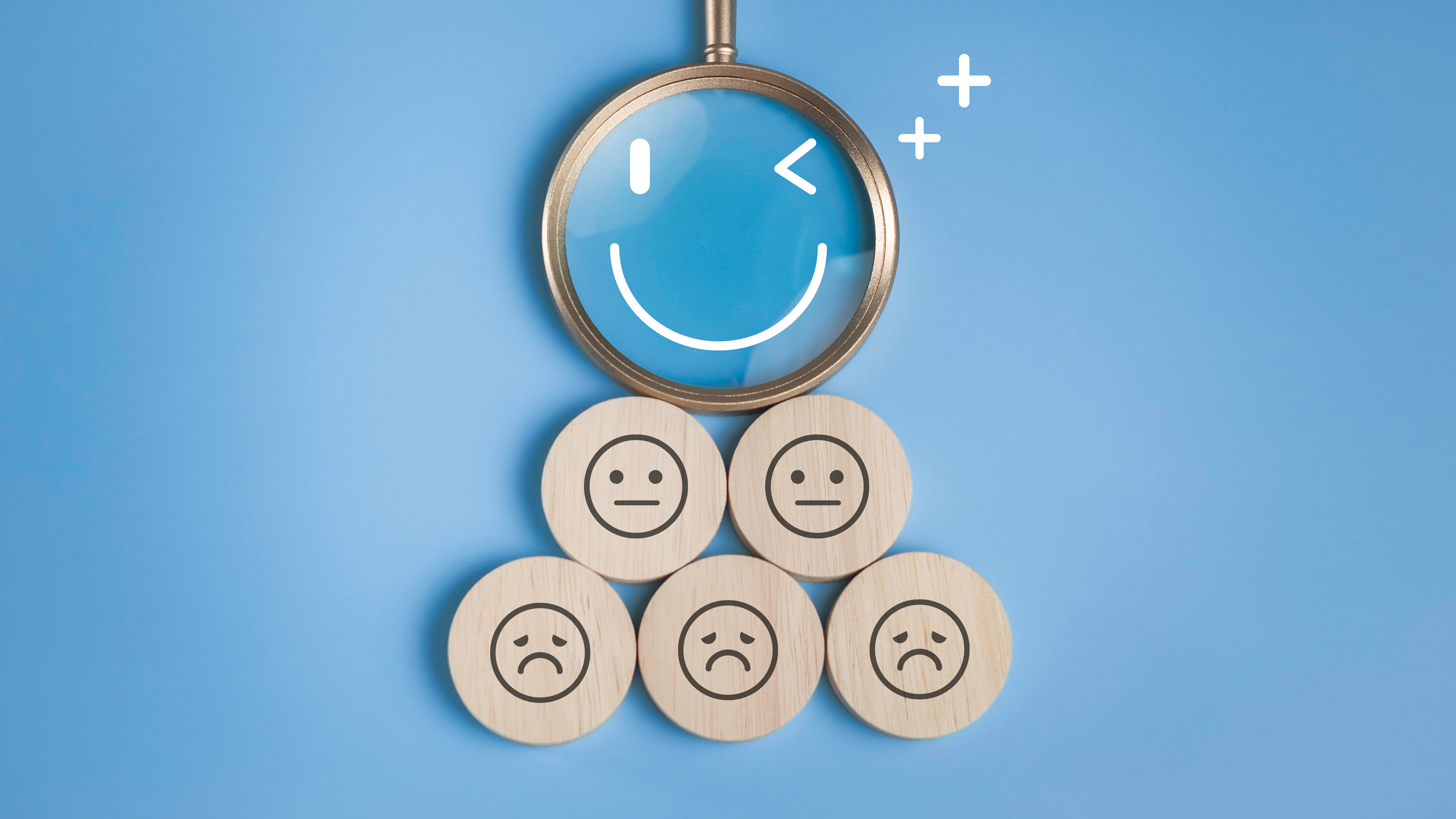 A magnifying glass focuses on a smiley face above three wooden tokens with sad and neutral faces on a blue background.