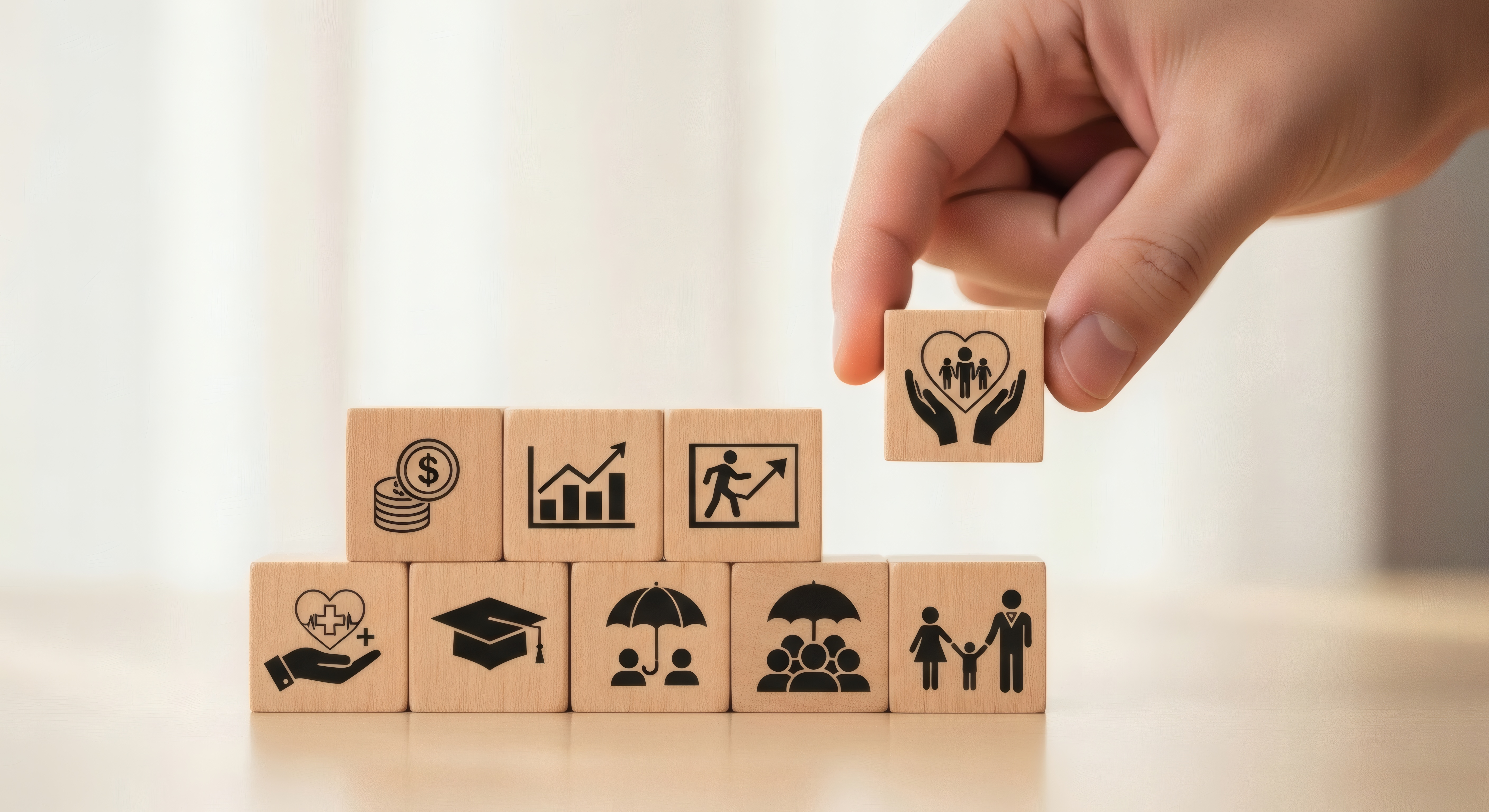 A hand places a block with a heart and family symbol atop a tower of wooden blocks depicting various icons like money and education.