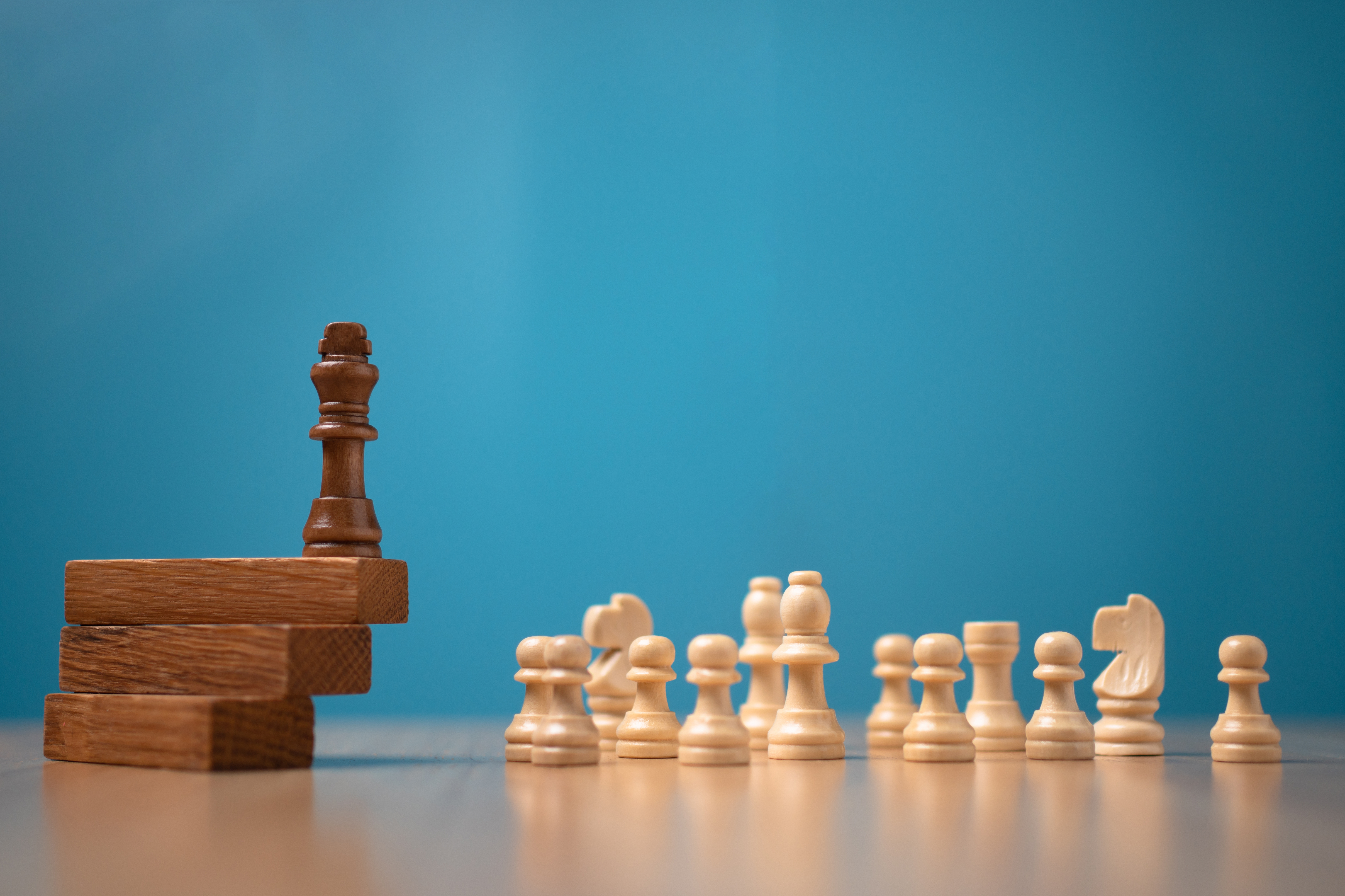 A wooden king chess piece stands on stacked blocks, facing a row of cream-colored chess pieces against a blue background.