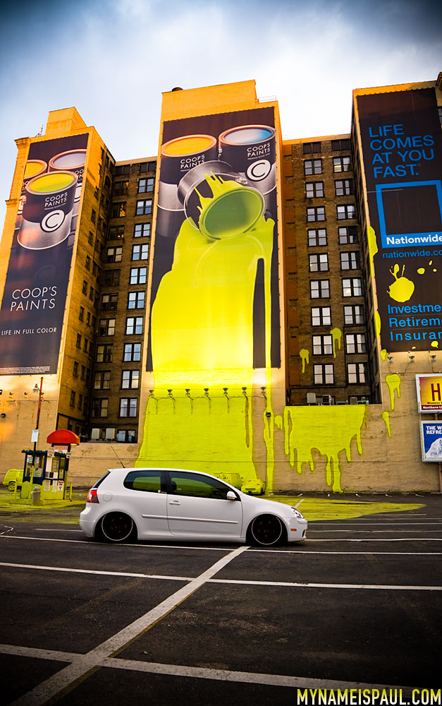 An image depicting a building and a parking lot. The building has advertisement posters for Coop's Paints and Nationwise Insurance. One of the Coop's Paints ads depicts a yellow paint bucket on it's side, spilling yellow paint past the poster's edge, onto the building wall and into the parking lot. Yellow splots can also be seen on the Nationwise Insurance ad poster. A white car in the parking lot is untouched, but another car in background of the picture appears to completely covered in yellow paint.