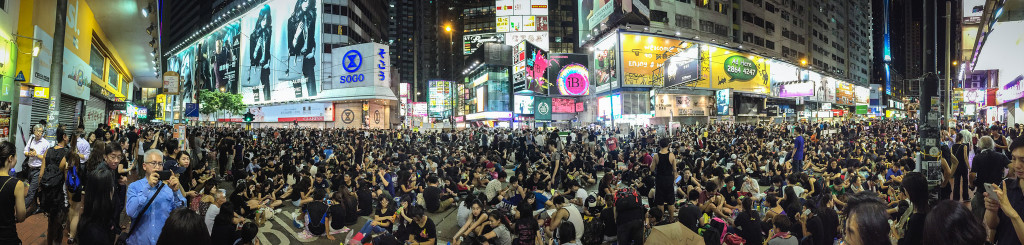 Panoramic color photo of busy Hong Kong intersection (not unlike Time Square in NYC) showing brightly lit digital ads and hundreds of people sitting in the street and on the sidewalks.