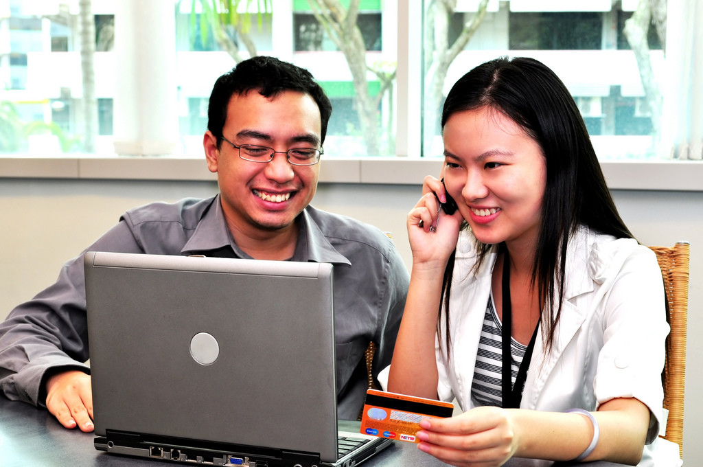 Foto de un hombre y una mujer mirando una computadora, sonriendo. La mujer está hablando por teléfono y sosteniendo una tarjeta de crédito.